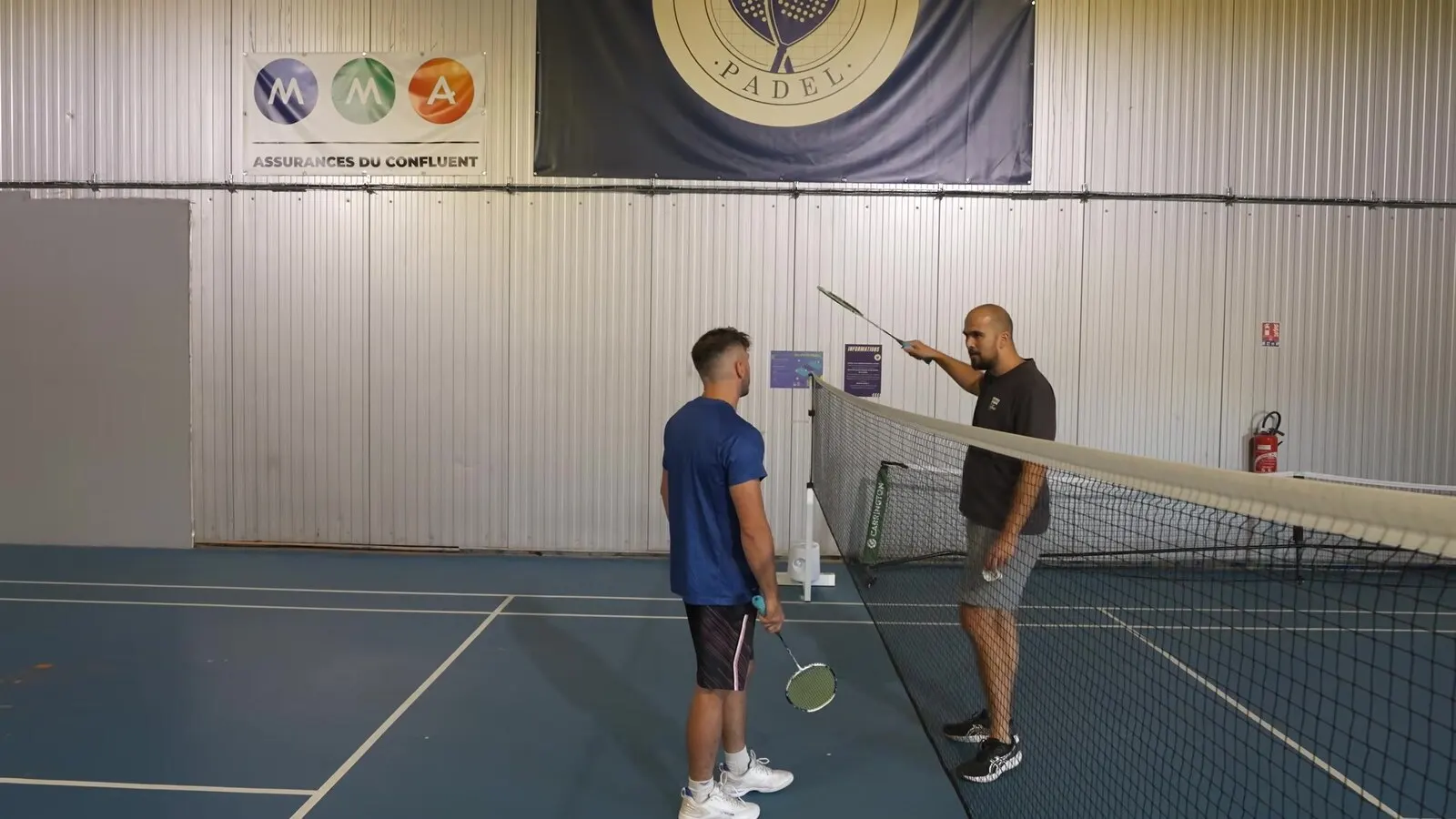 Au filet, un partenaire montre un geste de raquette à Richard Badminton pendant une séance d'entraînement