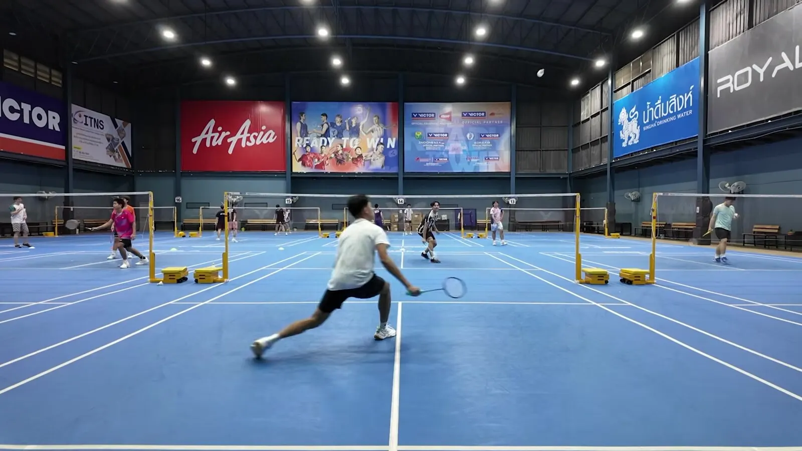 Joueur en fente profonde, bras tendu vers le volant, pendant un échange dans une grande salle de badminton