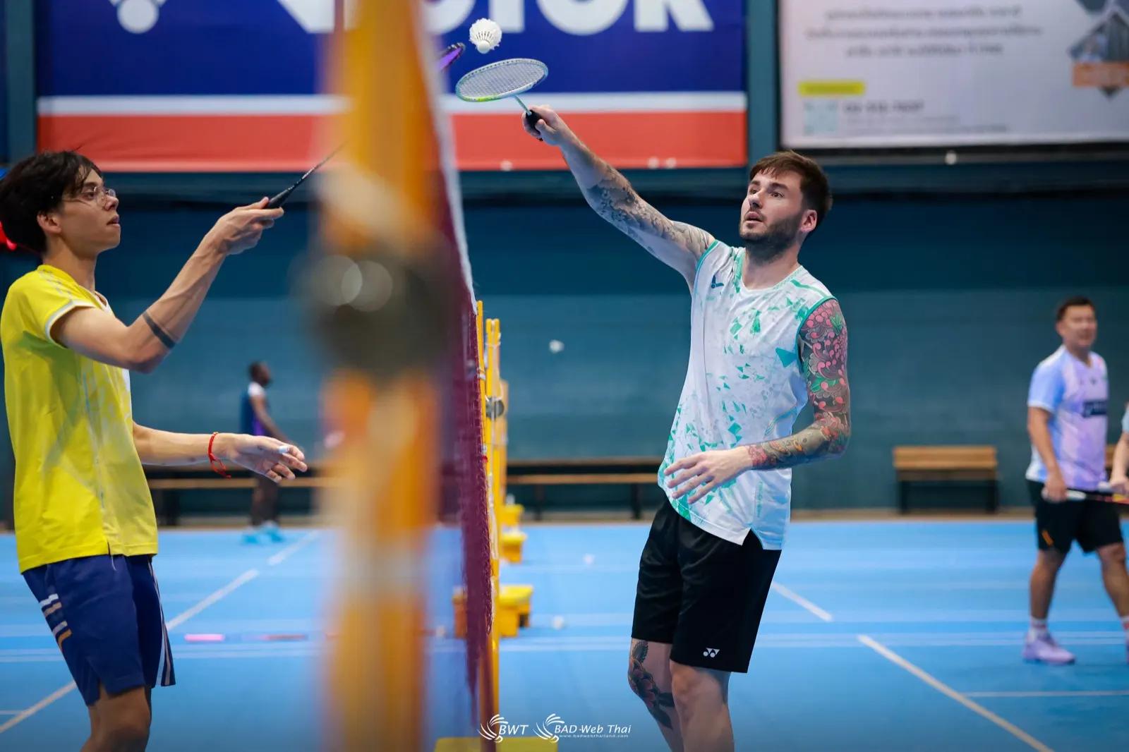 Duel au filet entre deux joueurs, raquettes hautes, pendant le stage de badminton à Bangkok